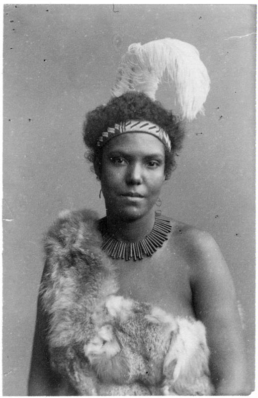Porträttfotografi av en ung svart kvinna, klädd i päls och med en stor vit fjäder i håret